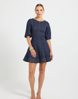 Woman wearing an indigo denim high rounded-neck mini dress with 3/4-length sleeves and elastic cuffs, complete with contrast top stitching running along the linear panels and wide hem.