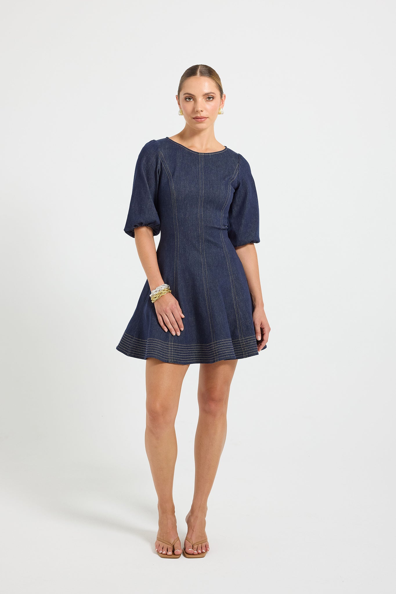 Woman wearing an indigo denim high rounded-neck mini dress with 3/4-length sleeves and elastic cuffs, complete with contrast top stitching running along the linear panels and wide hem.
