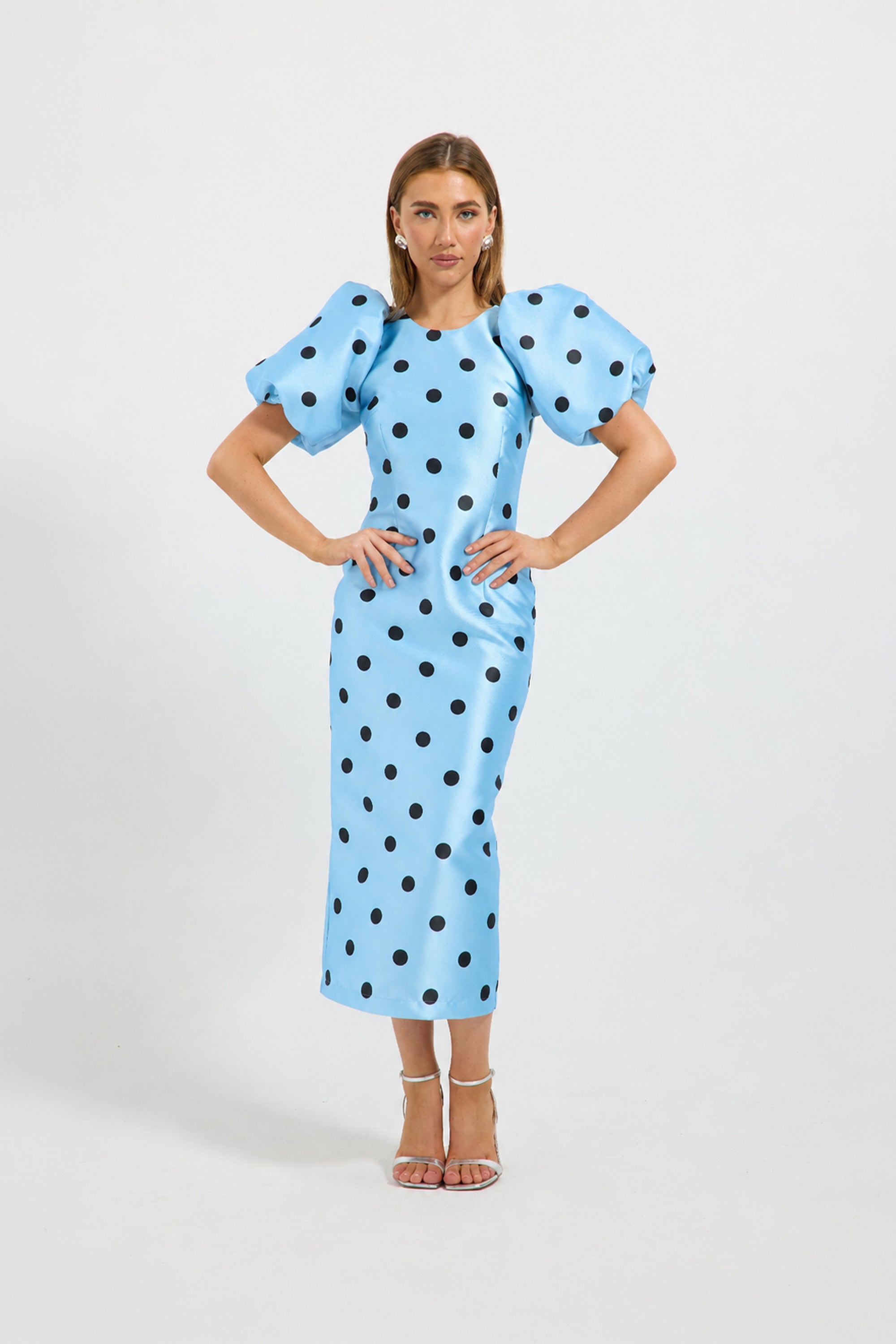 A woman modeling a light blue midi dress with black polka dots and dramatic puffed short sleeves, standing against a plain light gray studio background.