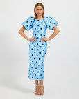 A woman modeling a light blue midi dress with black polka dots and dramatic puffed short sleeves, standing against a plain light gray studio background.