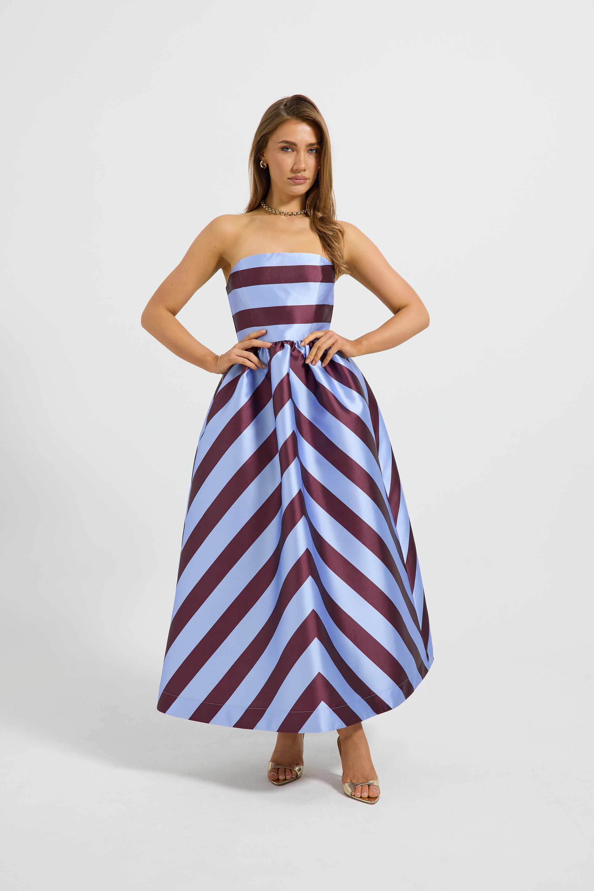 A woman modeling a strapless maxi dress with a chevron stripe in light blue and burgundy, standing against a light studio background.