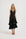 A blonde model wearing a sleeveless V halter neck black midi dress with a tiered ruffle skirt, photographed against a seamless white studio background.