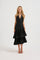 A blonde model wearing a sleeveless V halter neck black midi dress with a tiered ruffle skirt, photographed against a seamless white studio background.