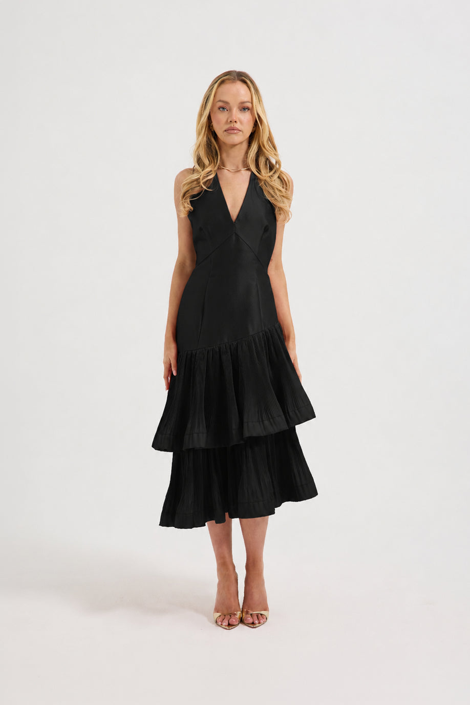 A blonde model wearing a sleeveless V halter neck black midi dress with a tiered ruffle skirt, photographed against a seamless white studio background.