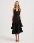A blonde model wearing a sleeveless V halter neck black midi dress with a tiered ruffle skirt, photographed against a seamless white studio background.