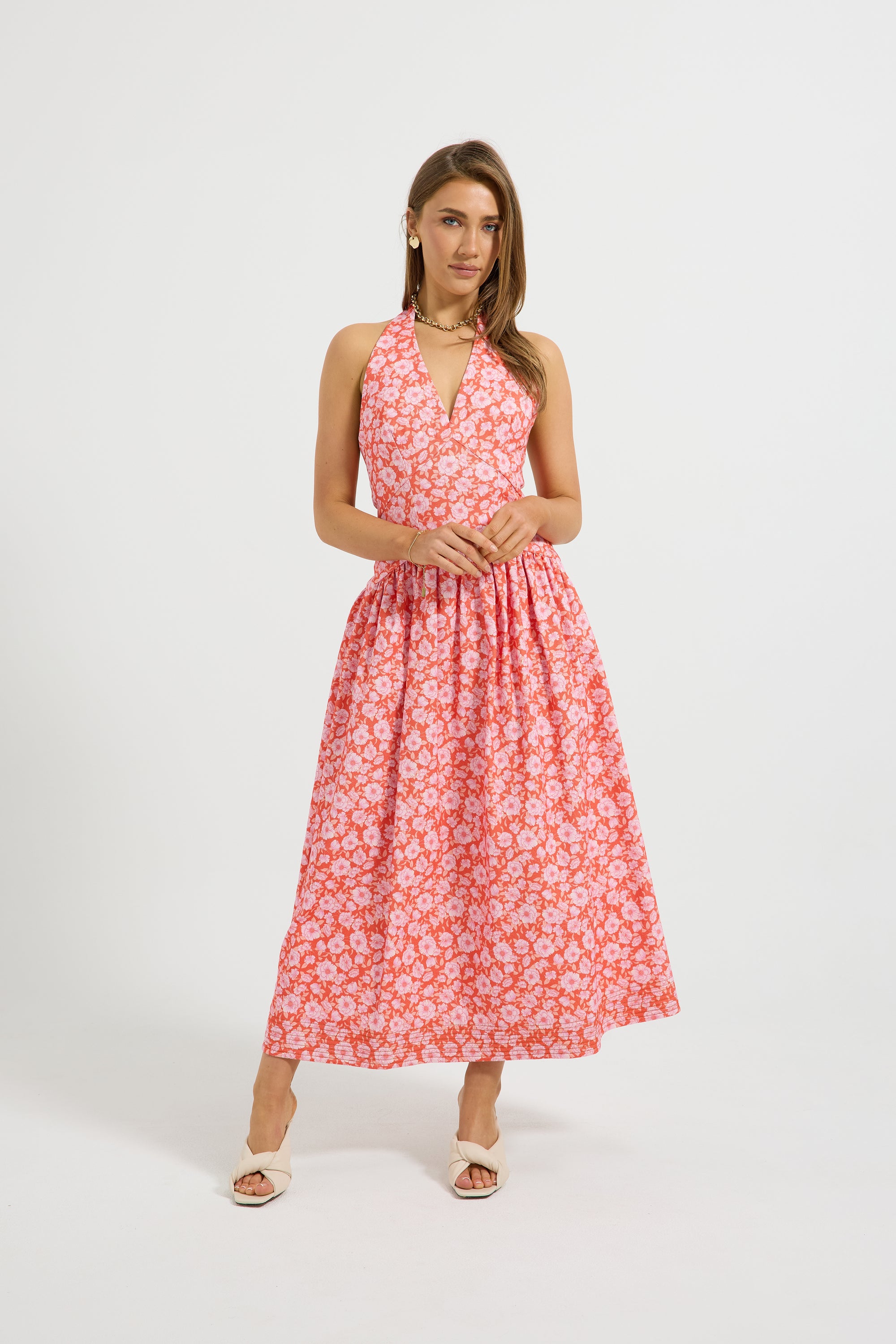 A woman modeling a sleeveless halter-neck midi dress with a white floral pattern on a coral pink background, paired with white slide sandals, set against a plain light gray studio background.