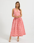 A woman modeling a sleeveless halter-neck midi dress with a white floral pattern on a coral pink background, paired with white slide sandals, set against a plain light gray studio background.