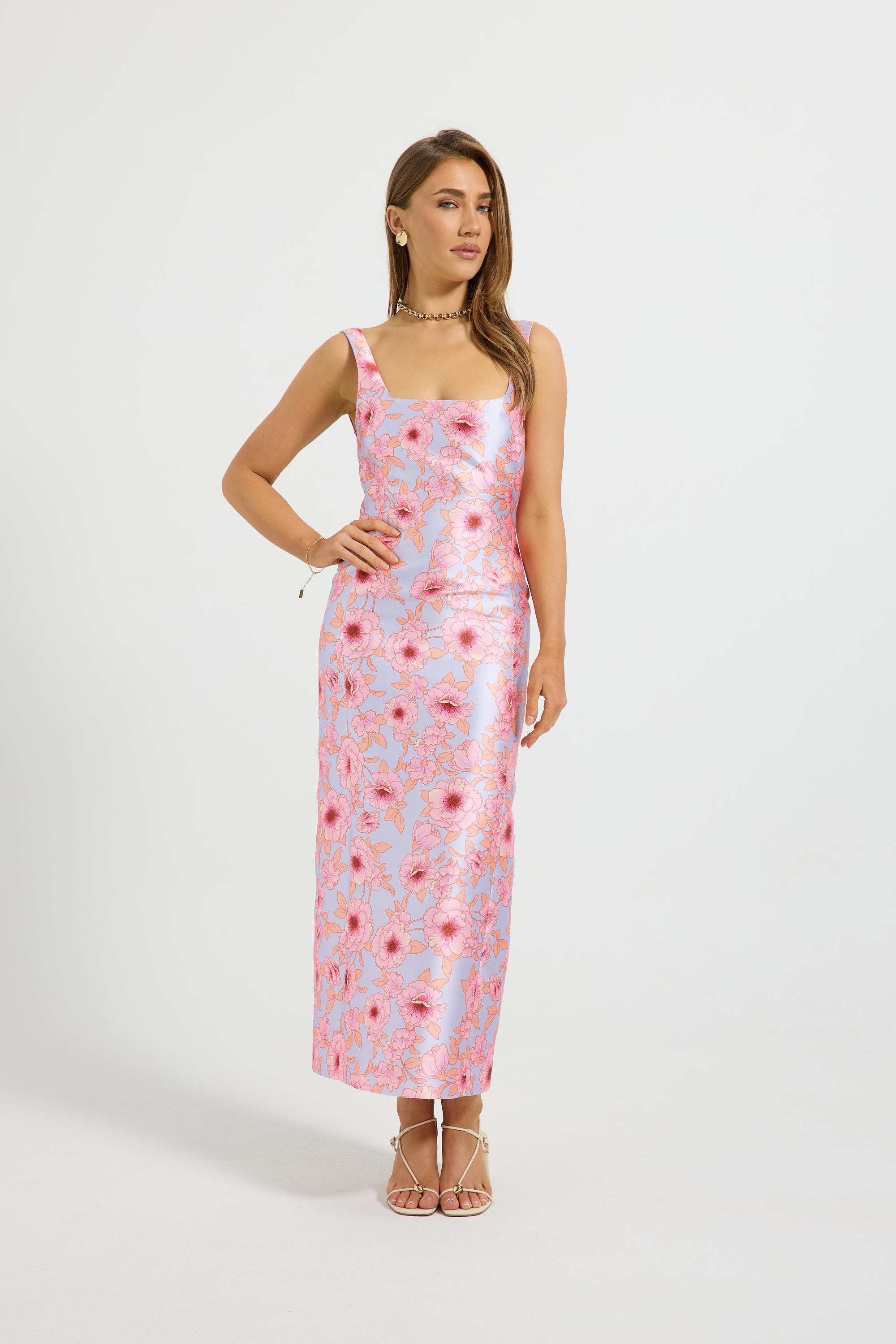 A woman modeling a sleeveless midi dress with a pink and light blue floral pattern, standing against a plain light gray studio background.