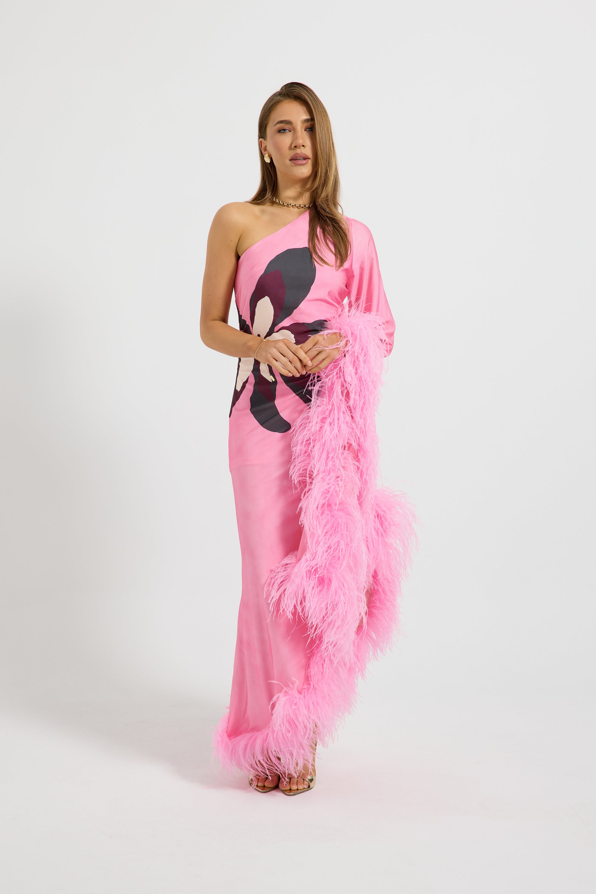 A woman models a one-shoulder pink gown with a black and white graphic print on the bodice and a fluffy pink feather boa draped over one arm, set against a plain light gray studio background.