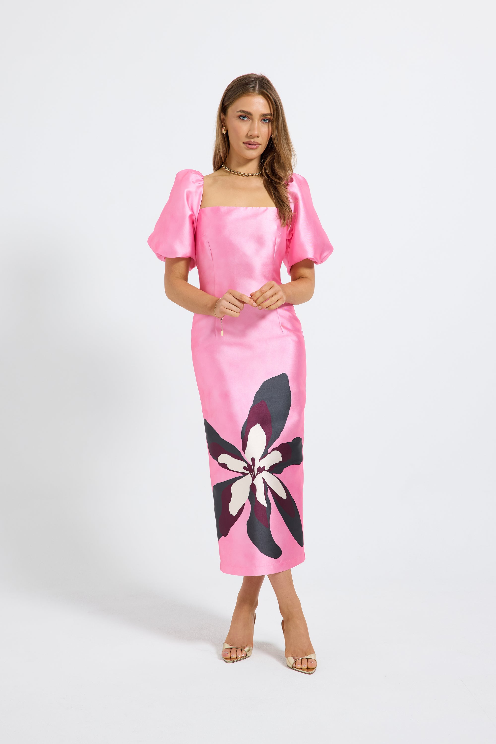 A woman modeling a bright pink midi dress with puff sleeves, a square neckline, and a large black and white floral print near the lower skirt, photographed against a light gray studio background.