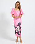 A woman modeling a bright pink midi dress with puff sleeves, a square neckline, and a large black and white floral print near the lower skirt, photographed against a light gray studio background.