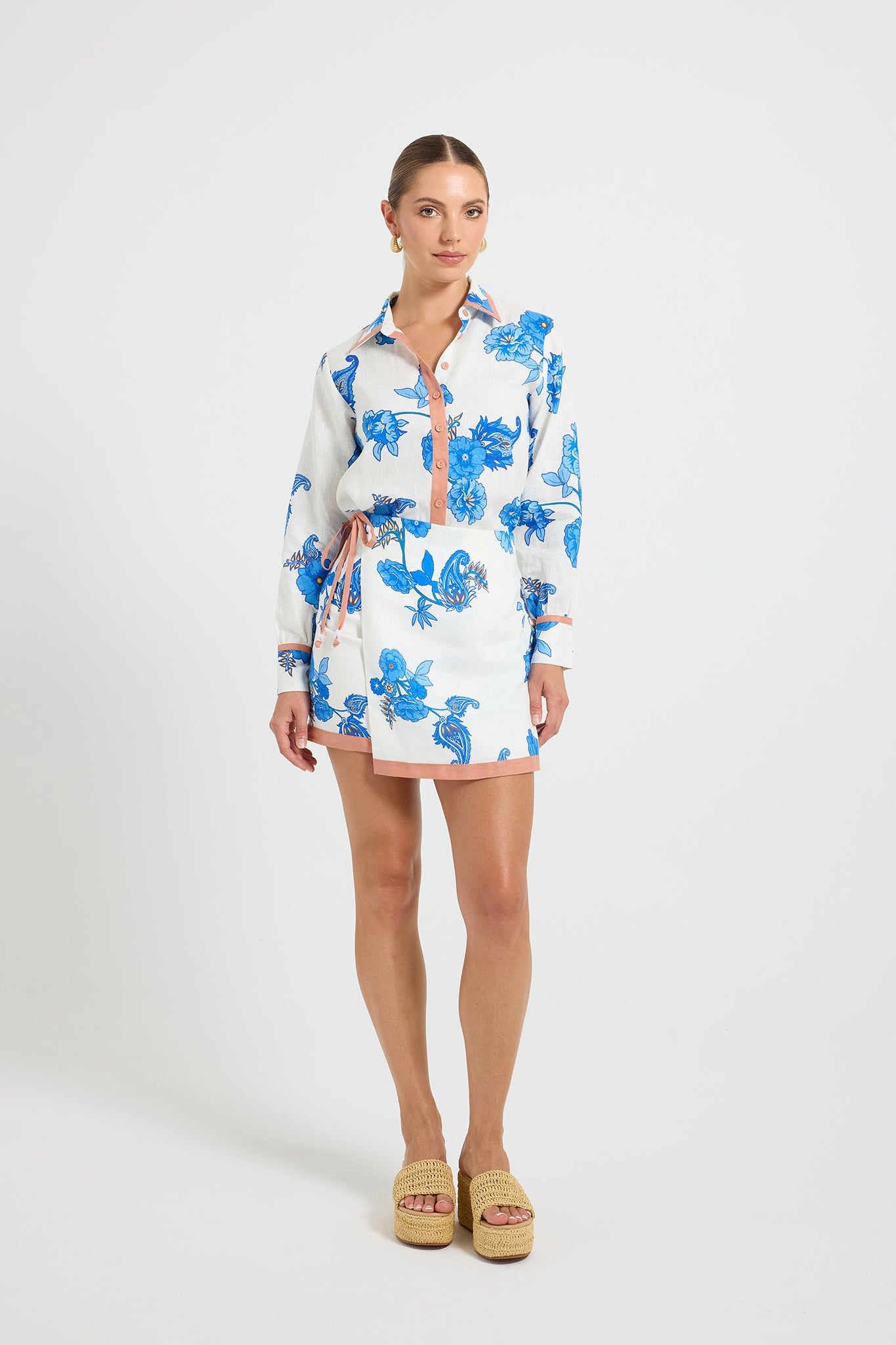 Woman wearing a white and blue floral print mini wrap skirt with tie detail and contrast binding, paired with a matching long-sleeve shirt.