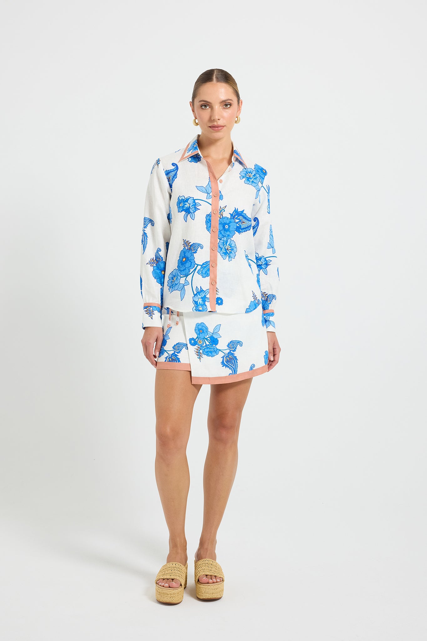 Woman wearing a white long sleeve button through shirt with blue floral pattern and matching skirt on a white background.