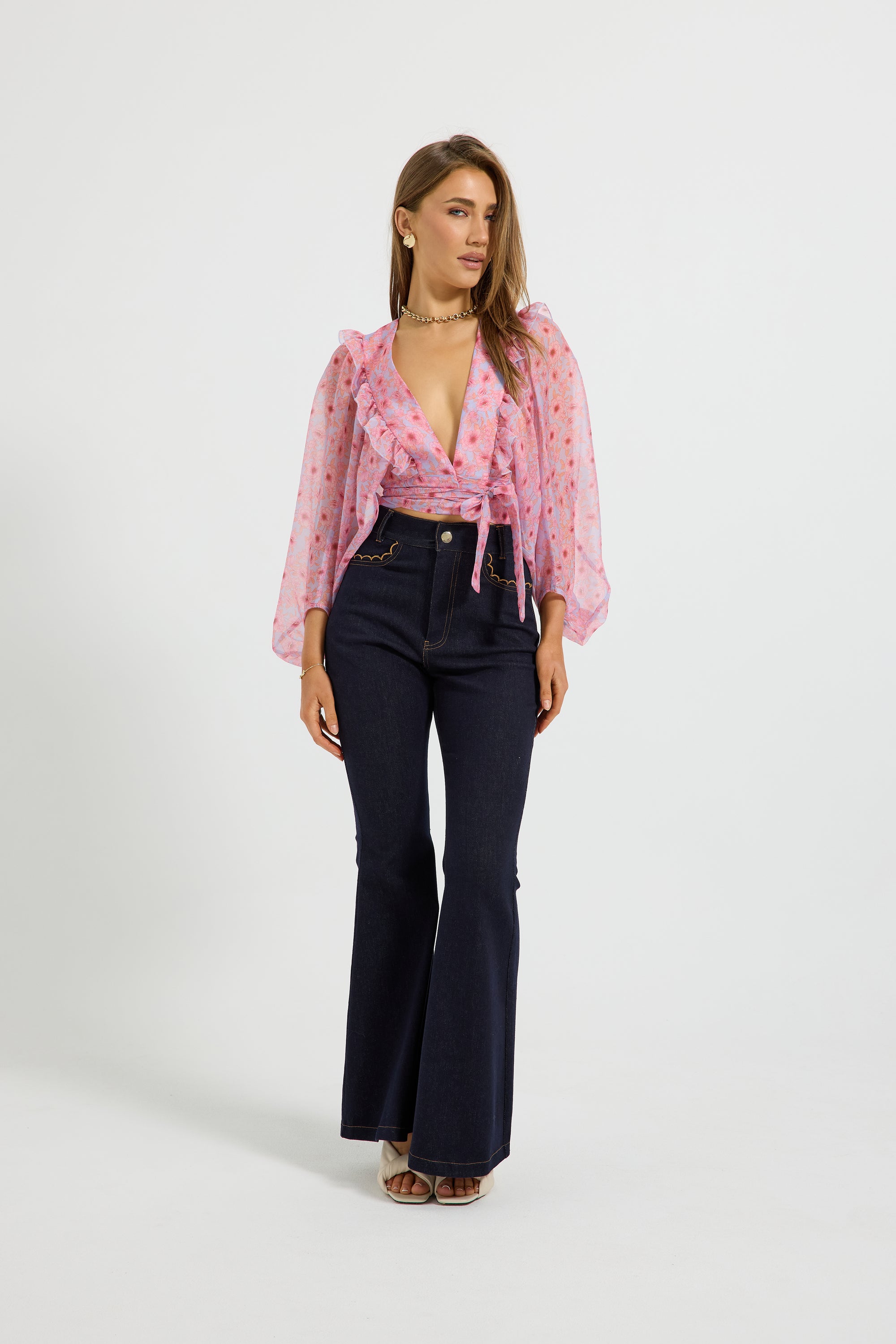 A woman modeling a pink floral long-sleeve tie-front blouse and dark navy flared jeans against a plain light gray studio background.